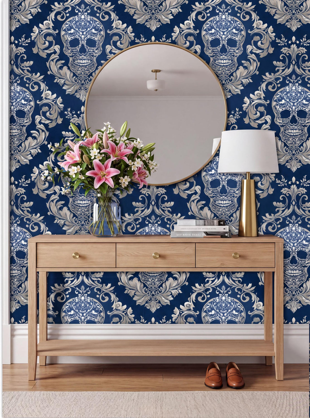 a wooden console table with a round mirror above it, set against a blue and white damask wallpaper with a skull design. The table has a vase of pink flowers and a lamp on it.