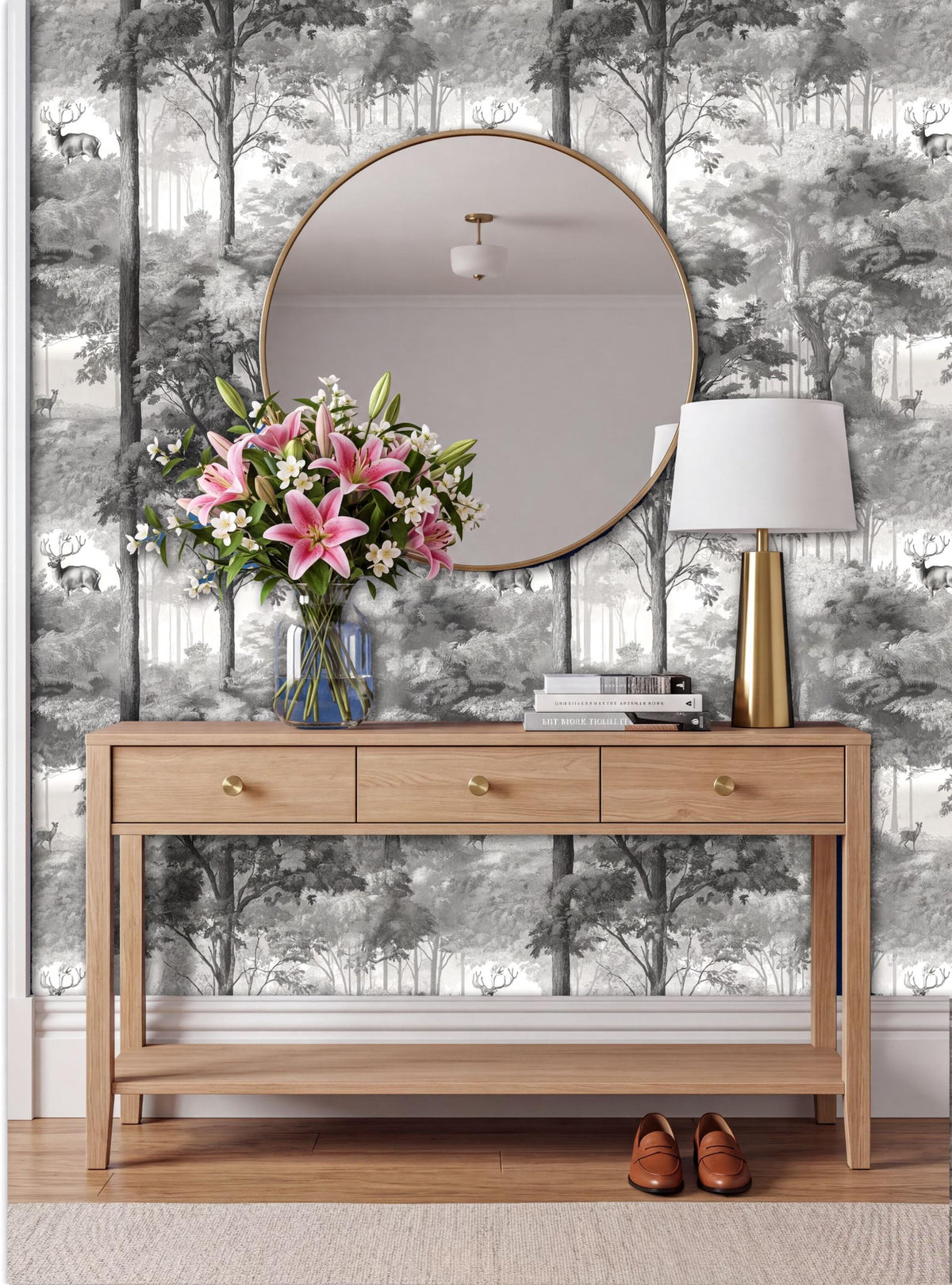 a wooden console table with a round mirror above it, a vase of pink lilies, and a lamp on the table. The table is set against a wall with a black and white wallpaper depicting a forest scene.