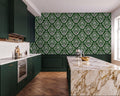 a modern kitchen with a large marble countertop and dark green cabinets. The walls are covered in a green and white damask pattern, creating a striking contrast with the marble countertop. The kitchen features a large island with a gold bowl on it, and a stainless steel oven is visible on the left side of the image.