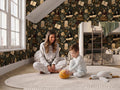 A woman and a young boy are sitting on the floor in a cozy bedroom, surrounded by stuffed animals and a large rug.