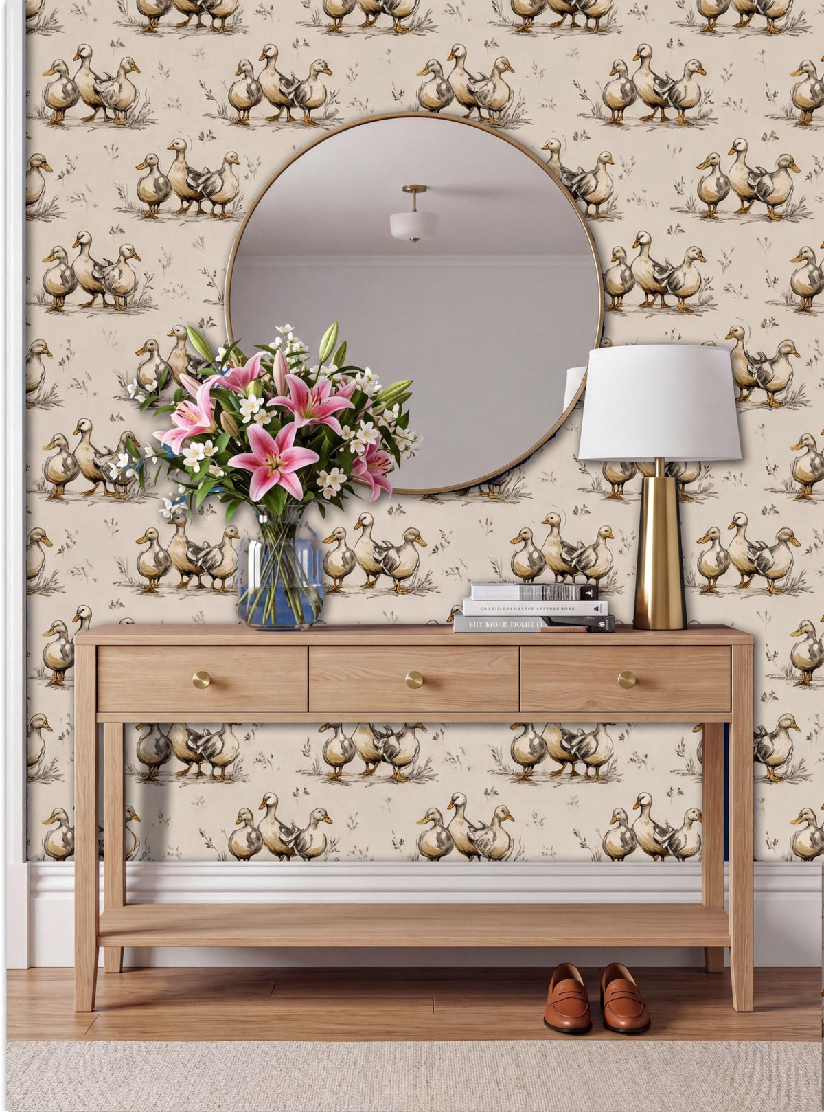 a wooden console table with a round mirror above it, a vase of pink flowers, and a lamp on the table. The wall behind the table has a repeating pattern of ducks.
