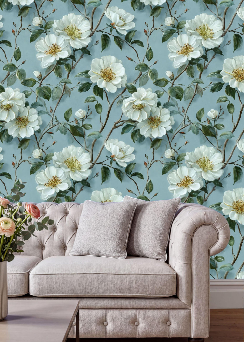a living room with a white tufted sofa and a vase of pink flowers on a coffee table. The sofa is positioned against a wall with a floral wallpaper pattern of white flowers and green leaves.