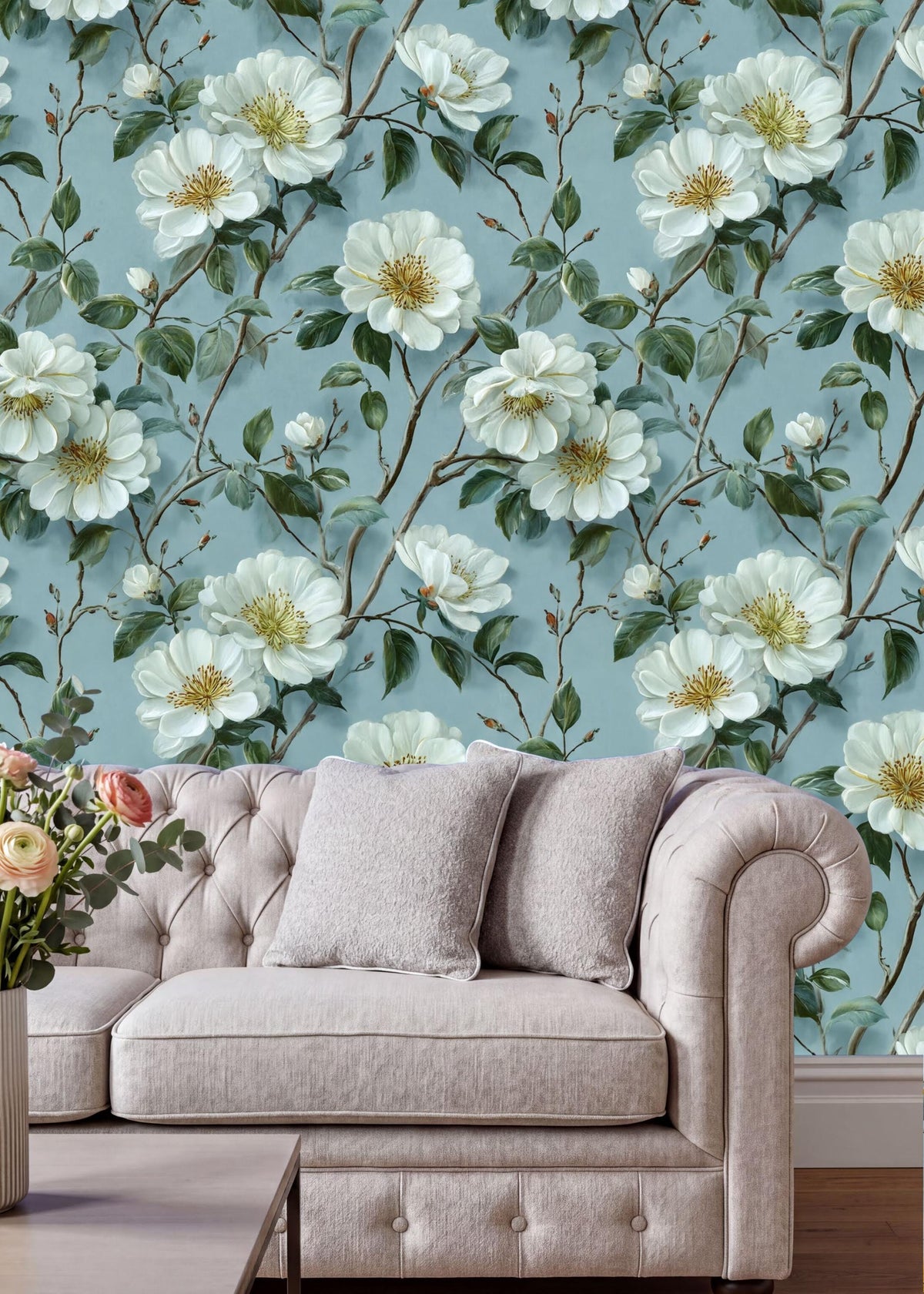 a living room with a white tufted sofa and a vase of pink flowers on a coffee table. The sofa is positioned against a wall with a floral wallpaper pattern of white flowers and green leaves.