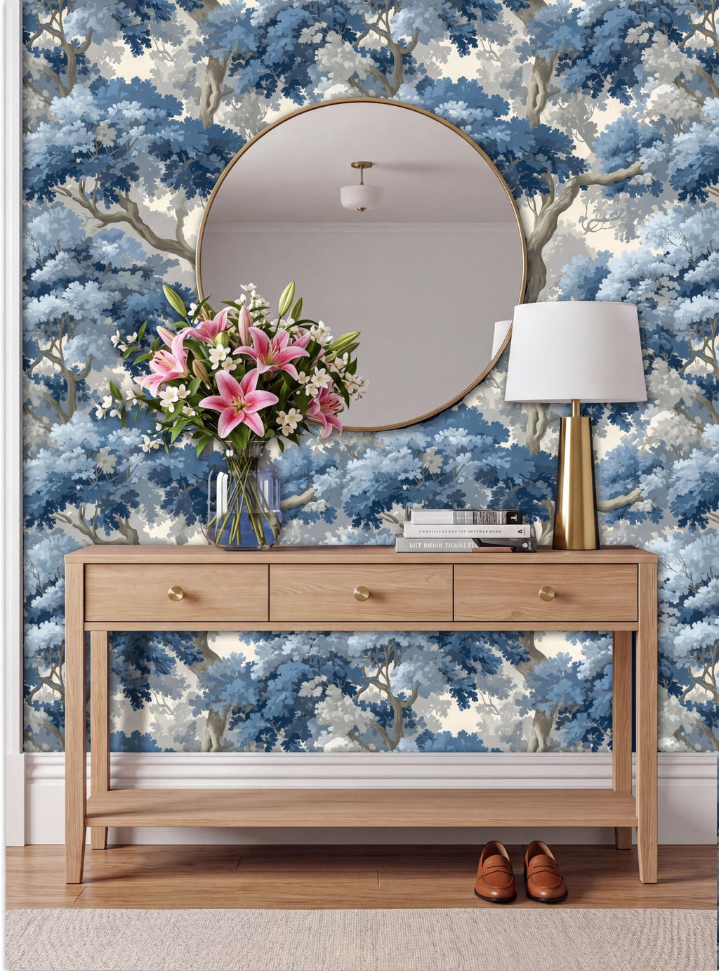 a wooden console table with a round mirror above it, set against a blue and white wallpaper with a forest scene. The table has three drawers and a vase of pink flowers. A lamp is also present on the table. A pair of shoes can be seen placed on the floor next to the table.