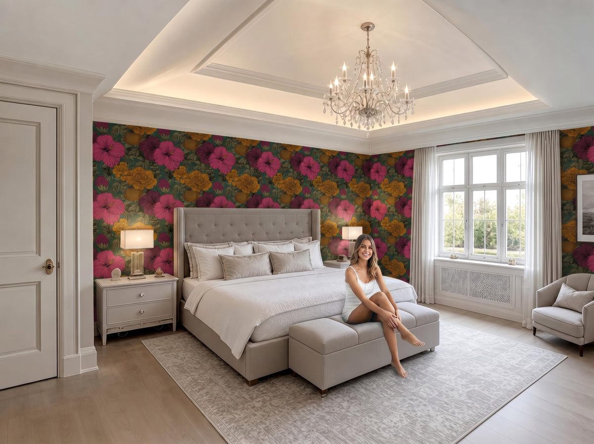 A woman sits on a gray ottoman in a luxurious bedroom with a large bed, floral wallpaper, and a chandelier overhead.
