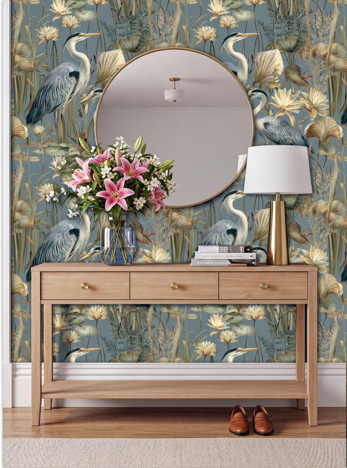 a wooden console table with a round mirror above it, set against a wall with a floral wallpaper. The table has a vase of pink flowers and a lamp on it.