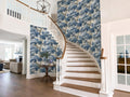 a spacious and elegant entryway with a wooden staircase, a wooden table, and a chandelier hanging from the ceiling. The walls are adorned with a blue and white wallpaper pattern, and the overall design has a harmonious and sophisticated aesthetic.