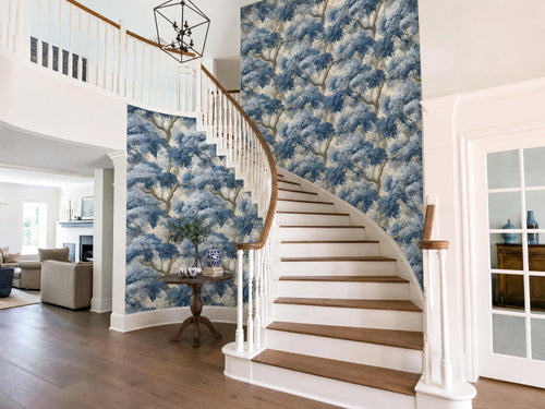 a spacious and elegant entryway with a wooden staircase, a wooden table, and a chandelier hanging from the ceiling. The walls are adorned with a blue and white wallpaper pattern, and the overall design has a harmonious and sophisticated aesthetic.