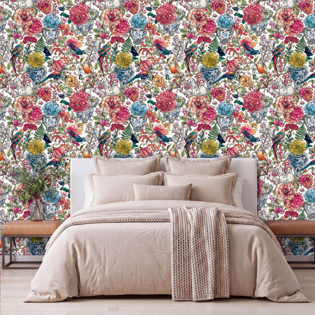 a bedroom with a large floral wallpaper behind a bed with a white headboard. The bed is dressed in a beige comforter and has a throw blanket draped over it. The room has a wooden nightstand on each side of the bed, and there is a vase of flowers on one of the nightstands.