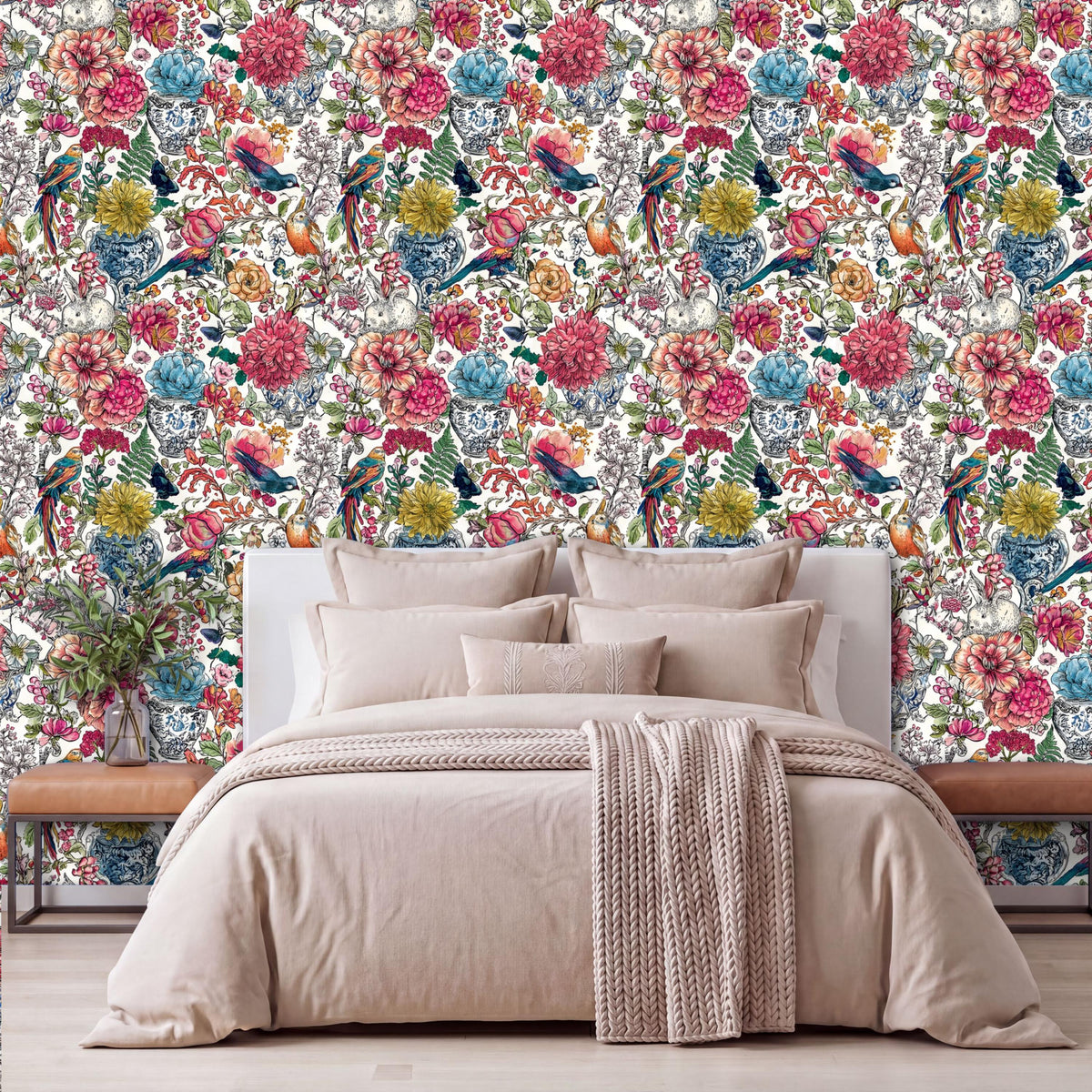 a bedroom with a large floral wallpaper behind a bed with a white headboard. The bed is dressed in a beige comforter and has a throw blanket draped over it. The room has a wooden nightstand on each side of the bed, and there is a vase of flowers on one of the nightstands.