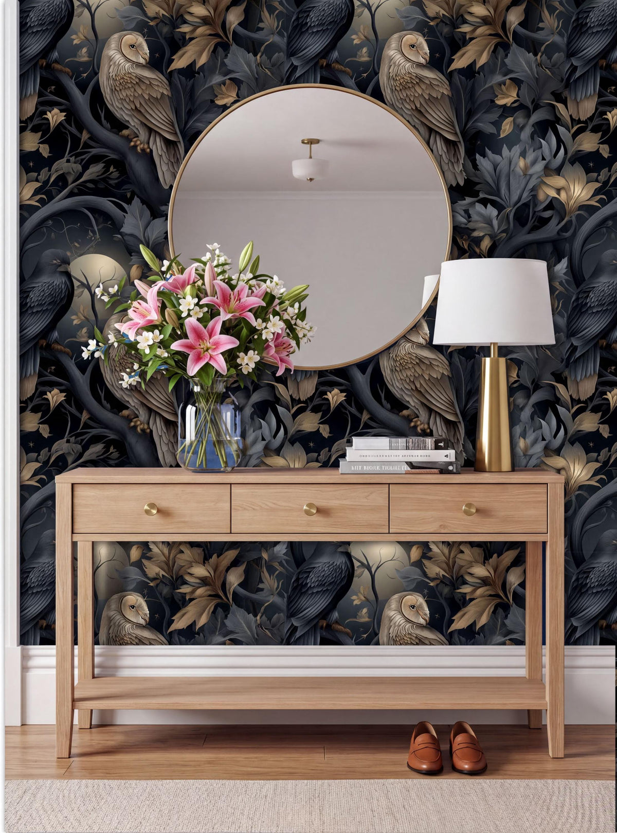 a wooden console table with a round mirror above it, a vase of pink flowers, a lamp, and a pair of brown shoes placed on the floor. The wall behind the table has a dark, ornate wallpaper with a repeating pattern of birds and leaves.