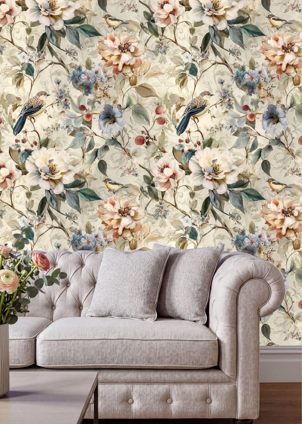 a cozy living room with a tufted cream-colored sofa adorned with two gray throw pillows. The sofa is positioned against a wall covered in a floral wallpaper, which adds a touch of nature and warmth to the space. In the background, there is a vase filled with flowers, further enhancing the room's inviting atmosphere.