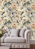 a cozy living room with a tufted cream-colored sofa adorned with two gray throw pillows. The sofa is positioned against a wall covered in a floral wallpaper, which adds a touch of nature and warmth to the space. In the background, there is a vase filled with flowers, further enhancing the room's inviting atmosphere.
