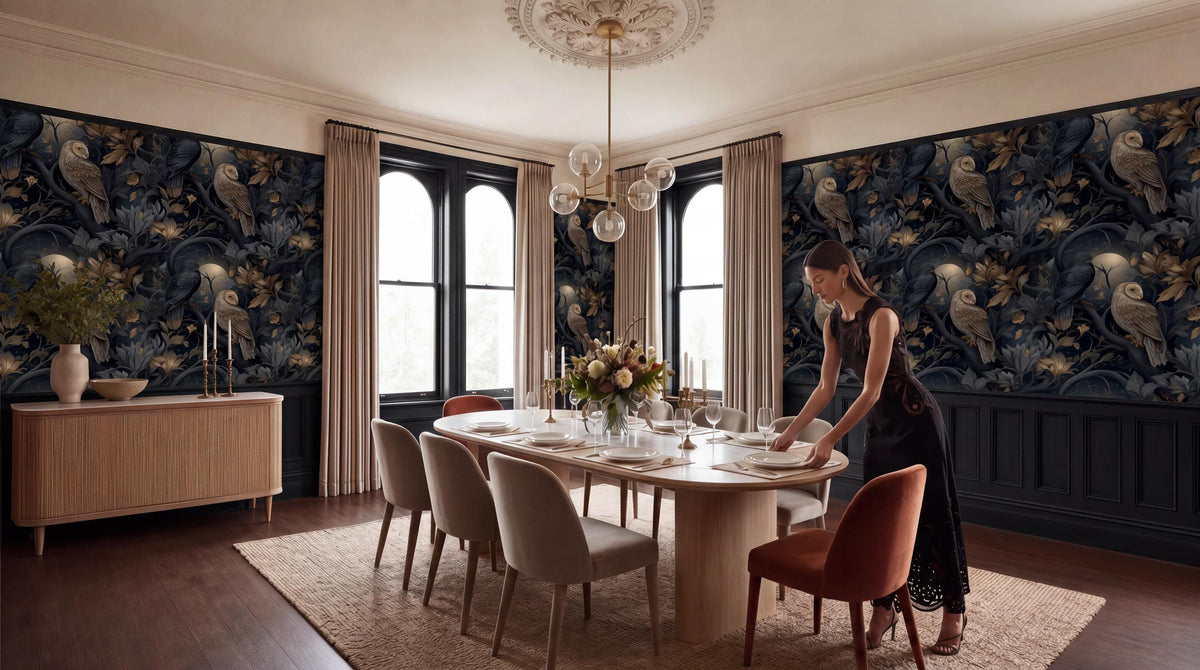 A woman in a black dress is setting the table in a dining room with a large floral wallpaper, a chandelier, and a wooden table with chairs.