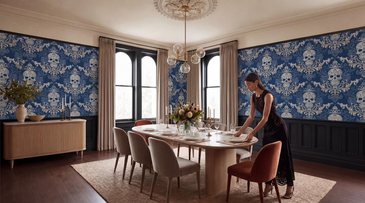 A woman in a black dress is setting the table in a dining room with a blue and white skull wallpaper, a chandelier, and a vase of flowers.