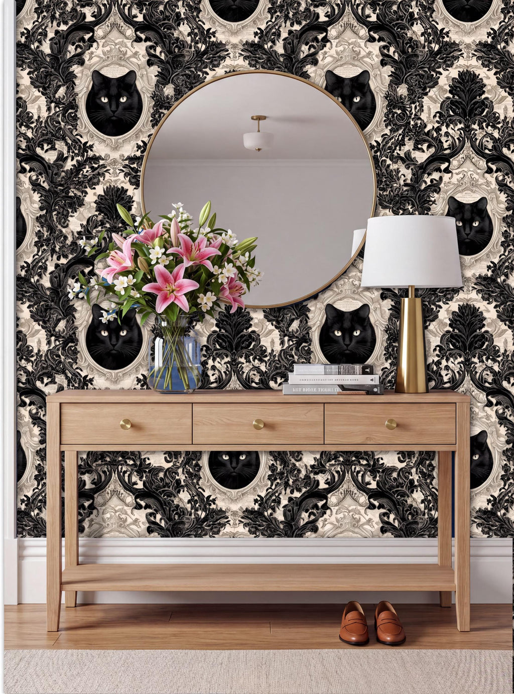 a wooden console table with a round mirror above it, set against a wall with a black and white damask pattern. The table has a vase of pink flowers and a lamp on it.