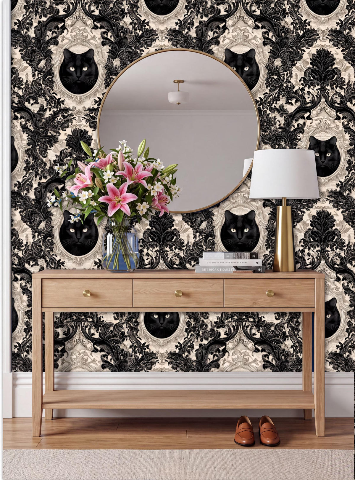 a wooden console table with a round mirror above it, set against a wall with a black and white damask pattern. The table has a vase of pink flowers and a lamp on it.