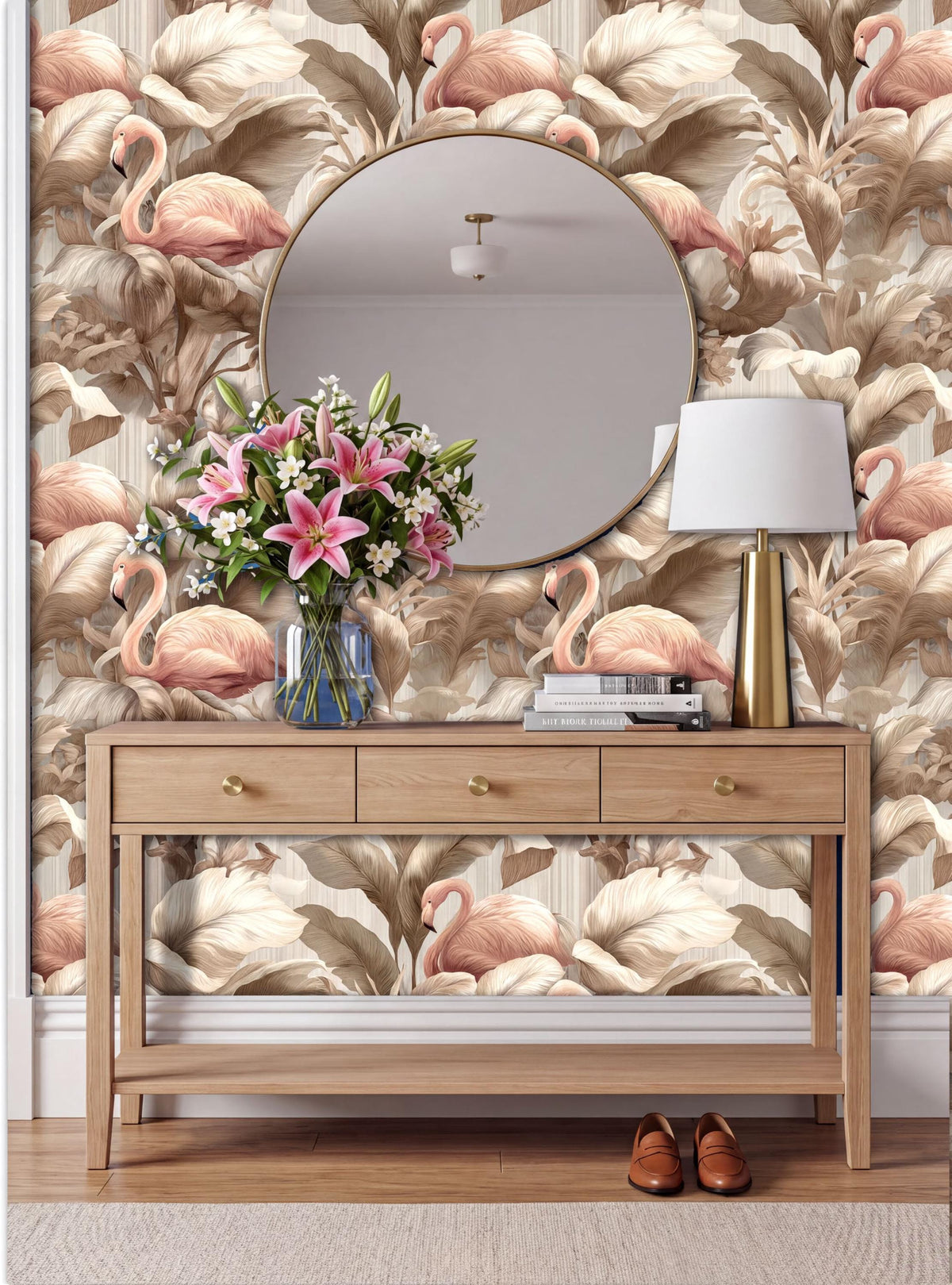 a wooden console table with a round mirror above it, a vase of pink flowers, a lamp, and a pair of brown shoes placed on the floor. The wall behind the table has a large floral wallpaper with pink flamingos and other tropical foliage.