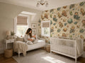 A woman sits on a white daybed in a nursery, holding a baby, with a crib and a wallpapered wall featuring a space-themed wallpaper.