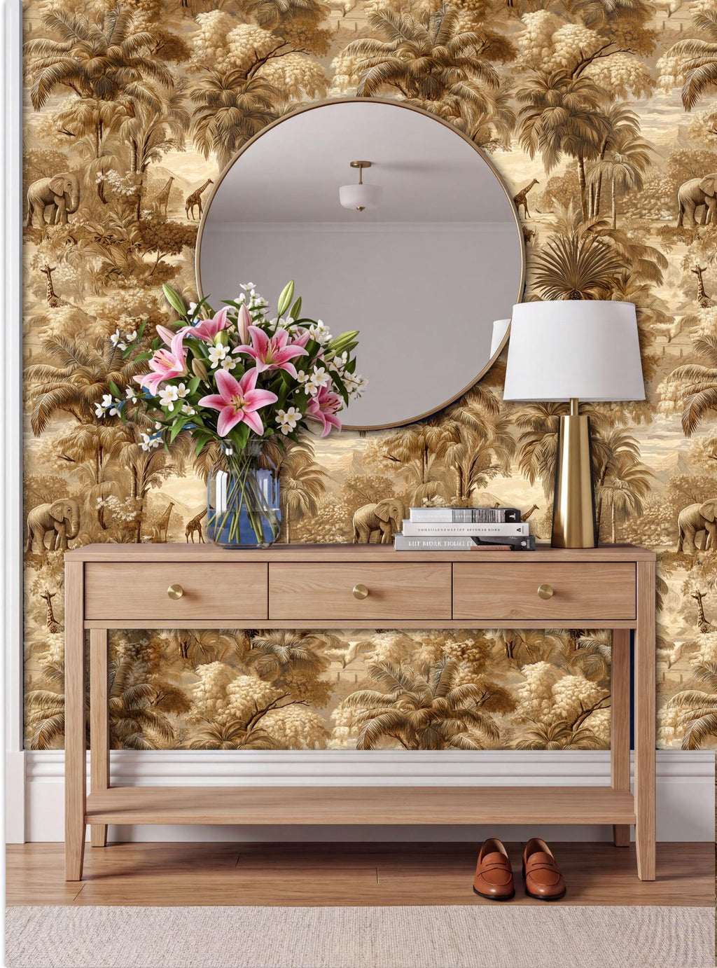 a wooden console table with a round mirror above it, a vase of pink lilies, a lamp, and a stack of books. The wall behind the table has a gold and brown wallpaper with palm trees and elephants.
