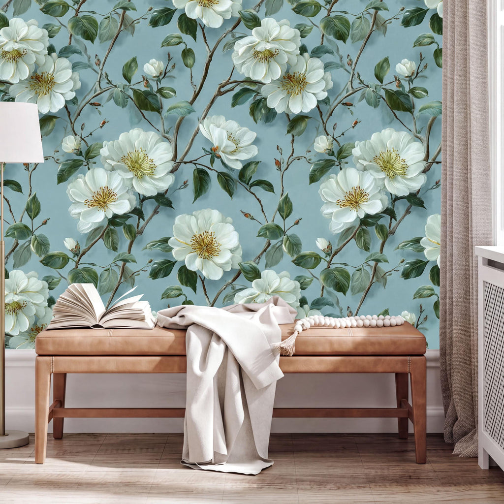 a wallpapered wall with a floral design of white flowers and green leaves, creating a serene and natural atmosphere. In the foreground, there is a wooden bench with an open book and a white blanket draped over it.