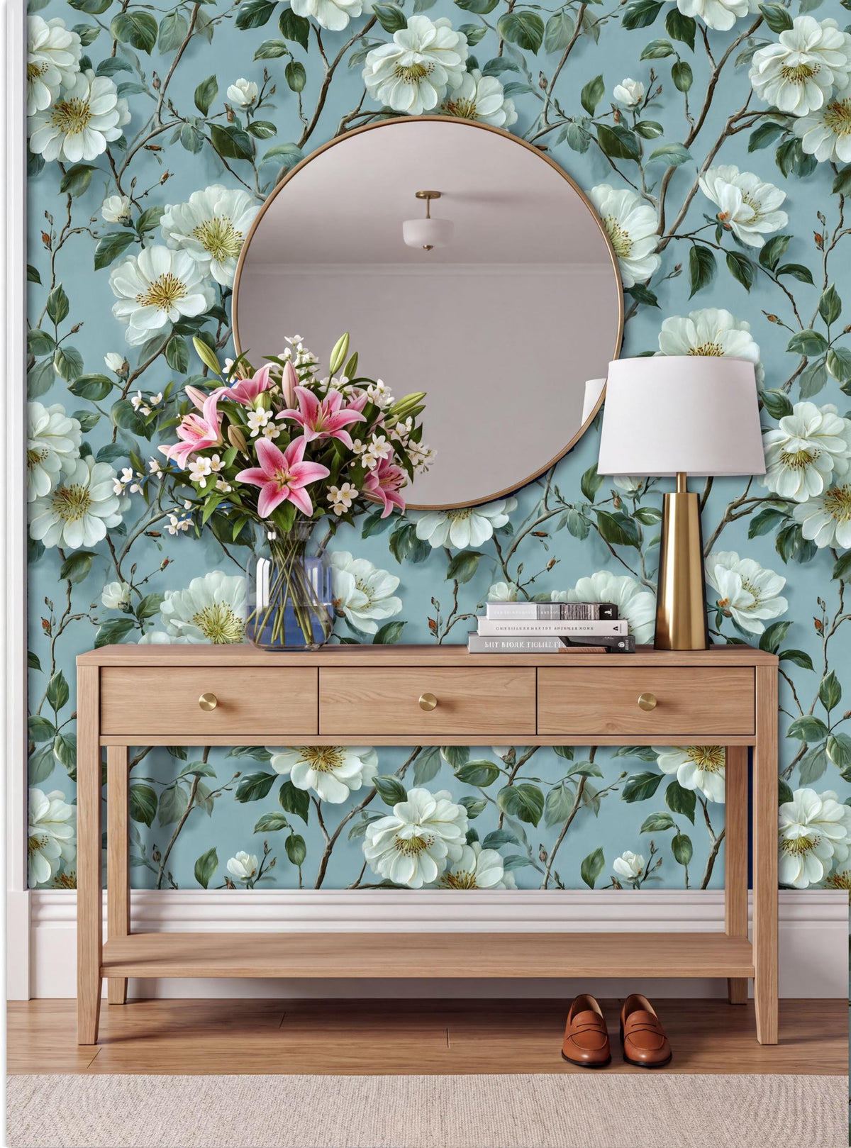 a wooden console table with a round mirror above it, a vase of pink lilies, a lamp, and a pair of brown shoes placed on the floor in front of the table.