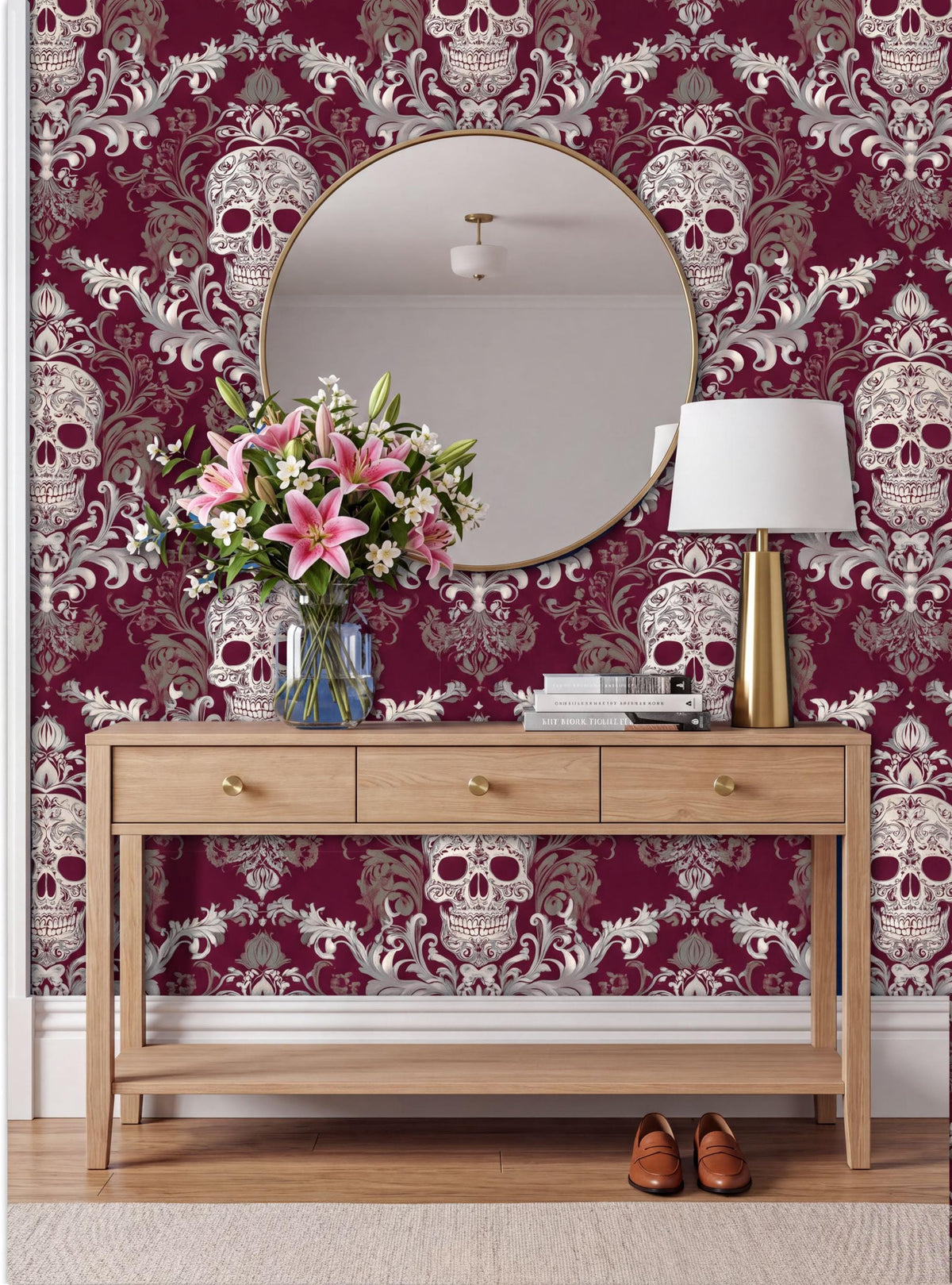 a wooden console table with a round mirror above it, set against a wall with a burgundy and white skull wallpaper. The table has a vase of pink flowers and a lamp on it.