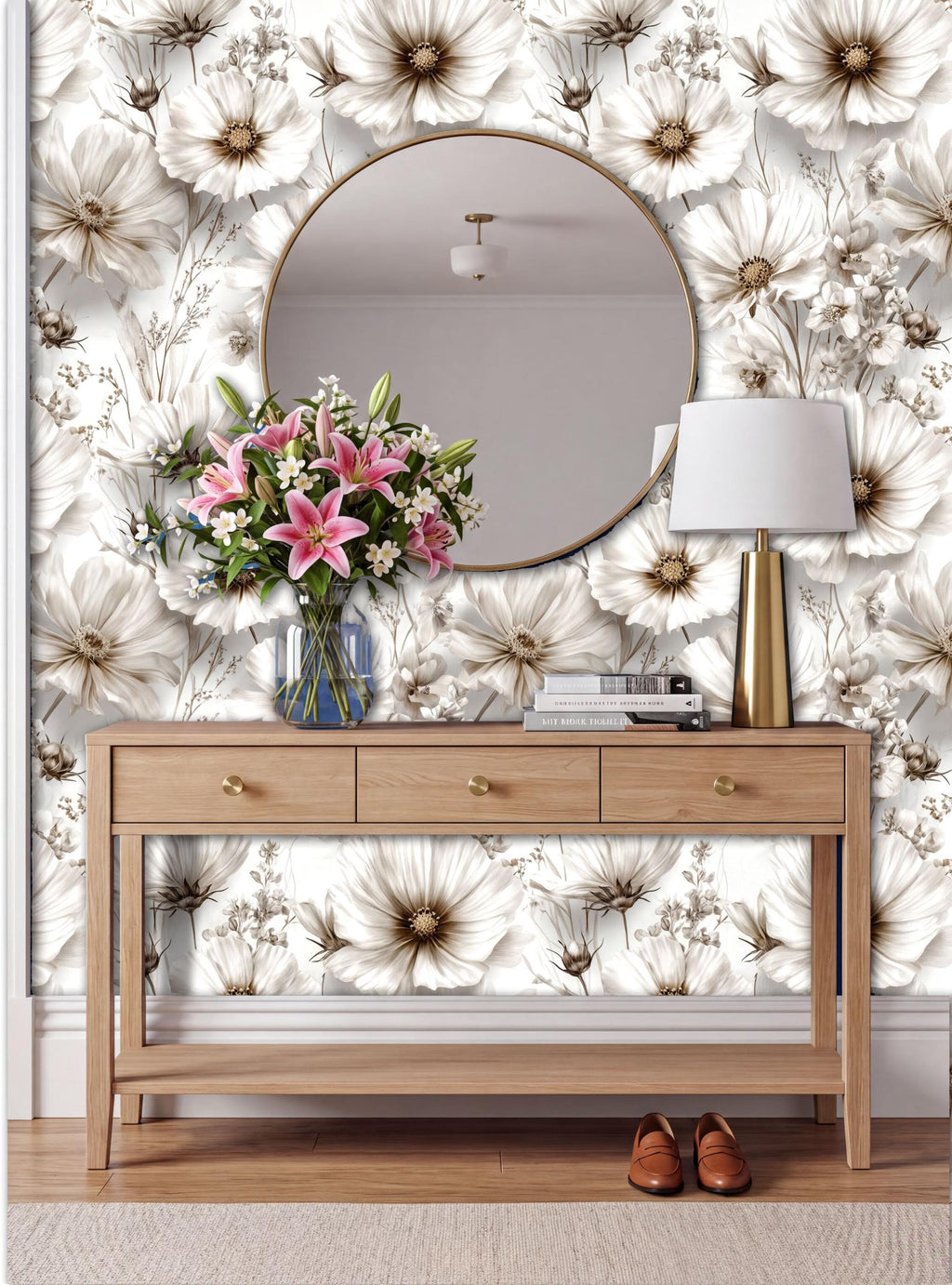 a wooden console table with a round mirror above it, set against a wall with a floral wallpaper. The table has a vase of pink and white flowers, and a lamp is placed on it. There are also some books on the table.