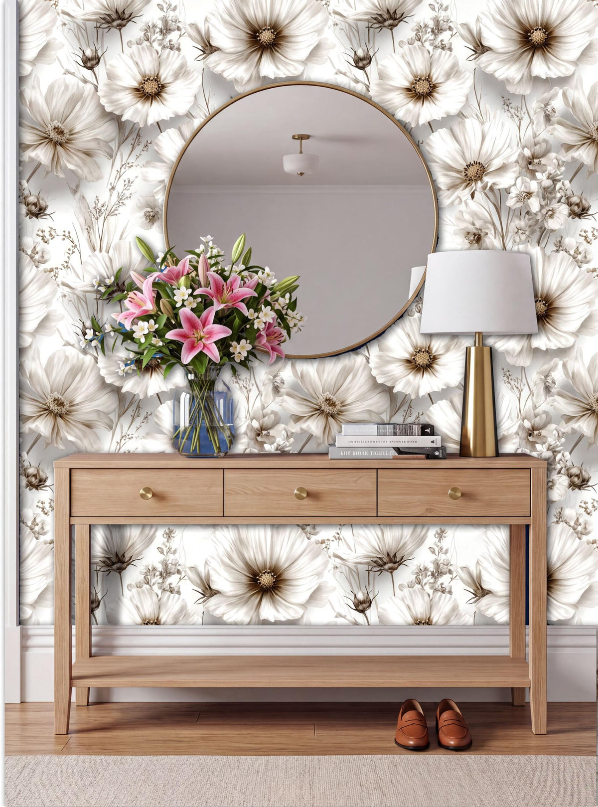 a wooden console table with a round mirror above it, set against a wall with a floral wallpaper. The table has a vase of pink and white flowers, and a lamp is placed on it. There are also some books on the table.