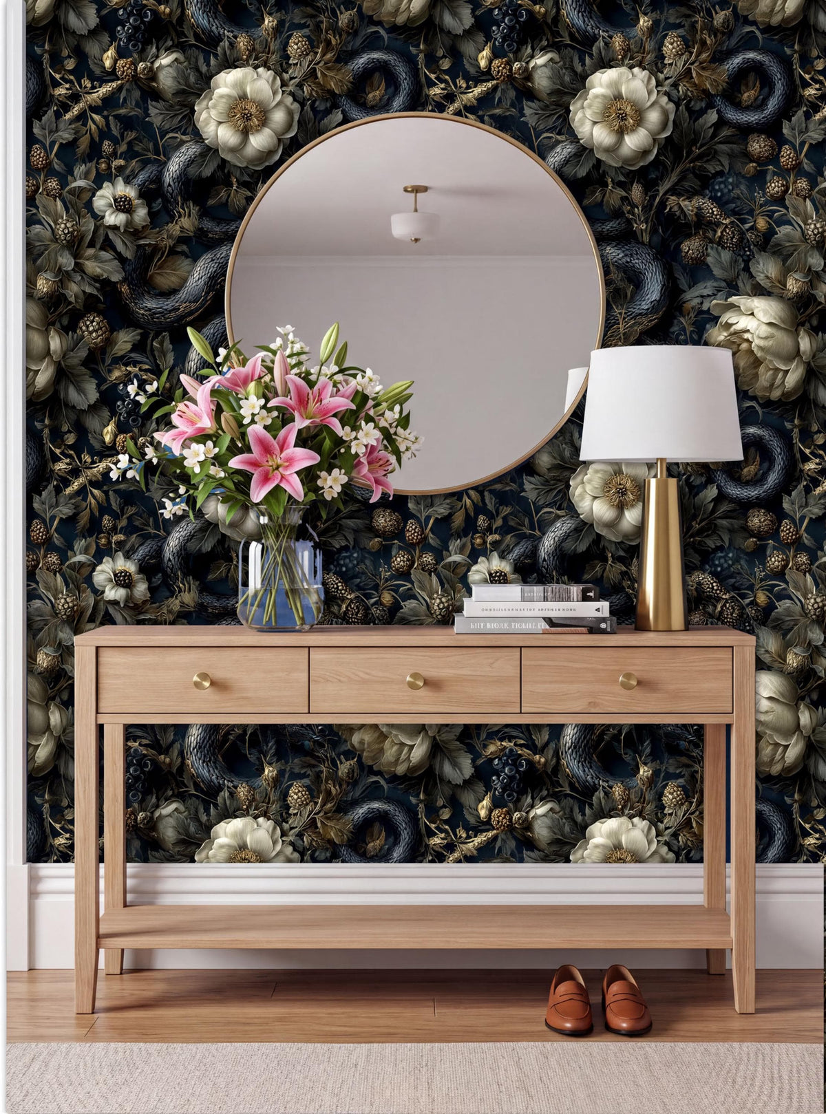 a wooden console table with a round mirror above it, a vase of pink lilies, a lamp, and a pair of brown shoes placed on a rug. The wall behind the table is covered in a dark floral wallpaper.