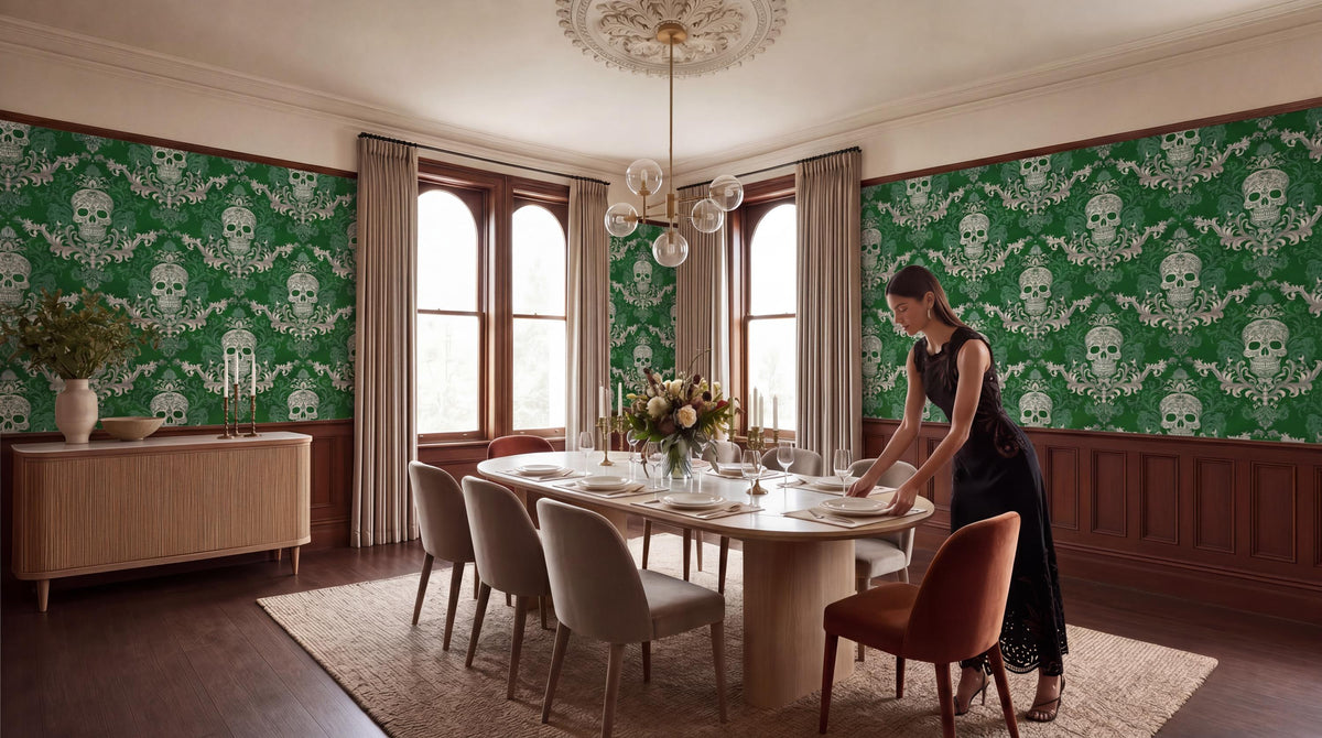 A woman in a black dress is setting the table in a dining room with a large, ornate chandelier overhead. The room has green and white skull wallpaper, a wooden dining table with white plates and silverware, and a large window with curtains.