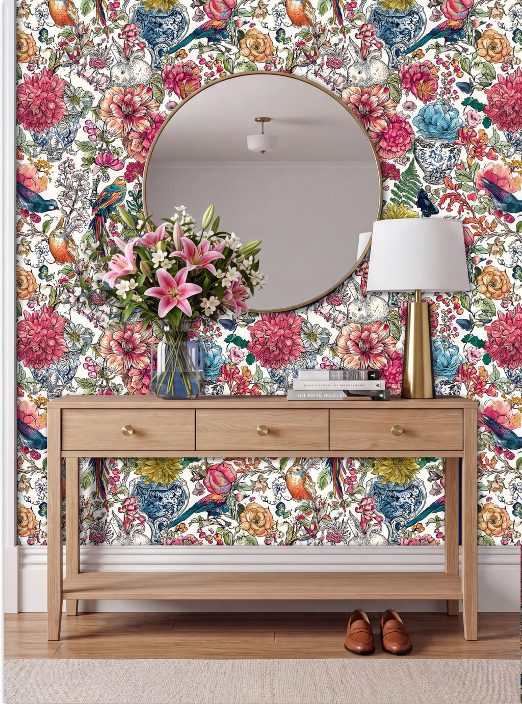 a wooden console table with a round mirror above it, set against a vibrant floral wallpaper. The table has three drawers and a vase of flowers on top. A lamp is also present on the table.