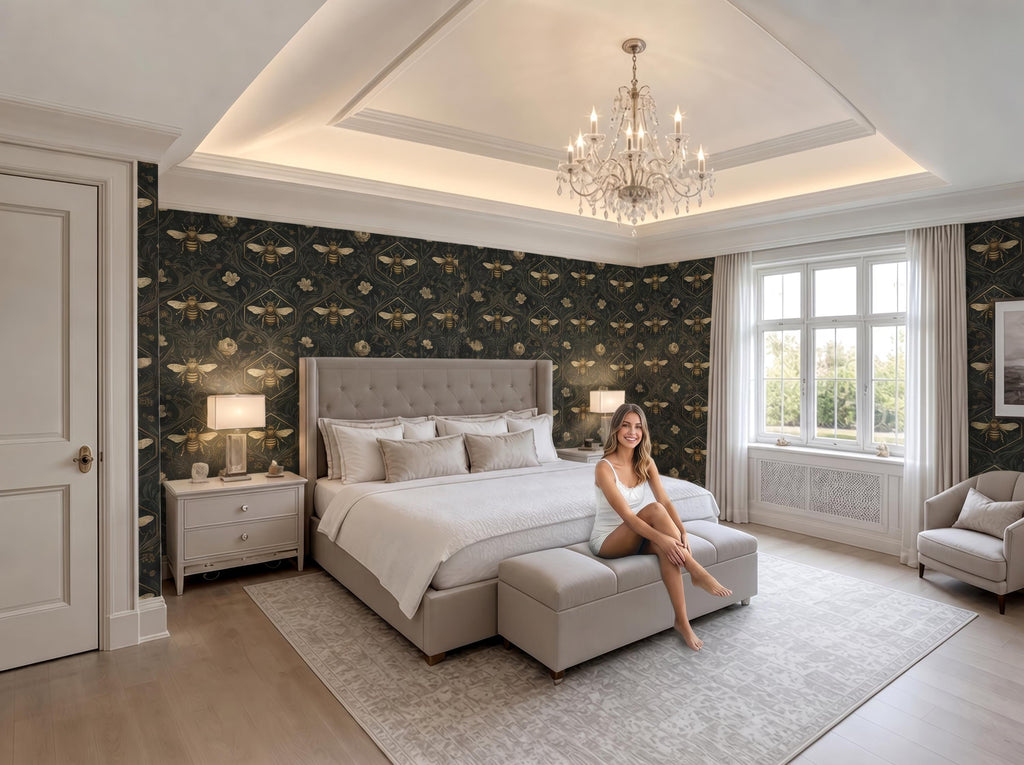 A woman sits on a bench in a luxurious bedroom with a large bed, a chandelier, and a patterned wallpaper.