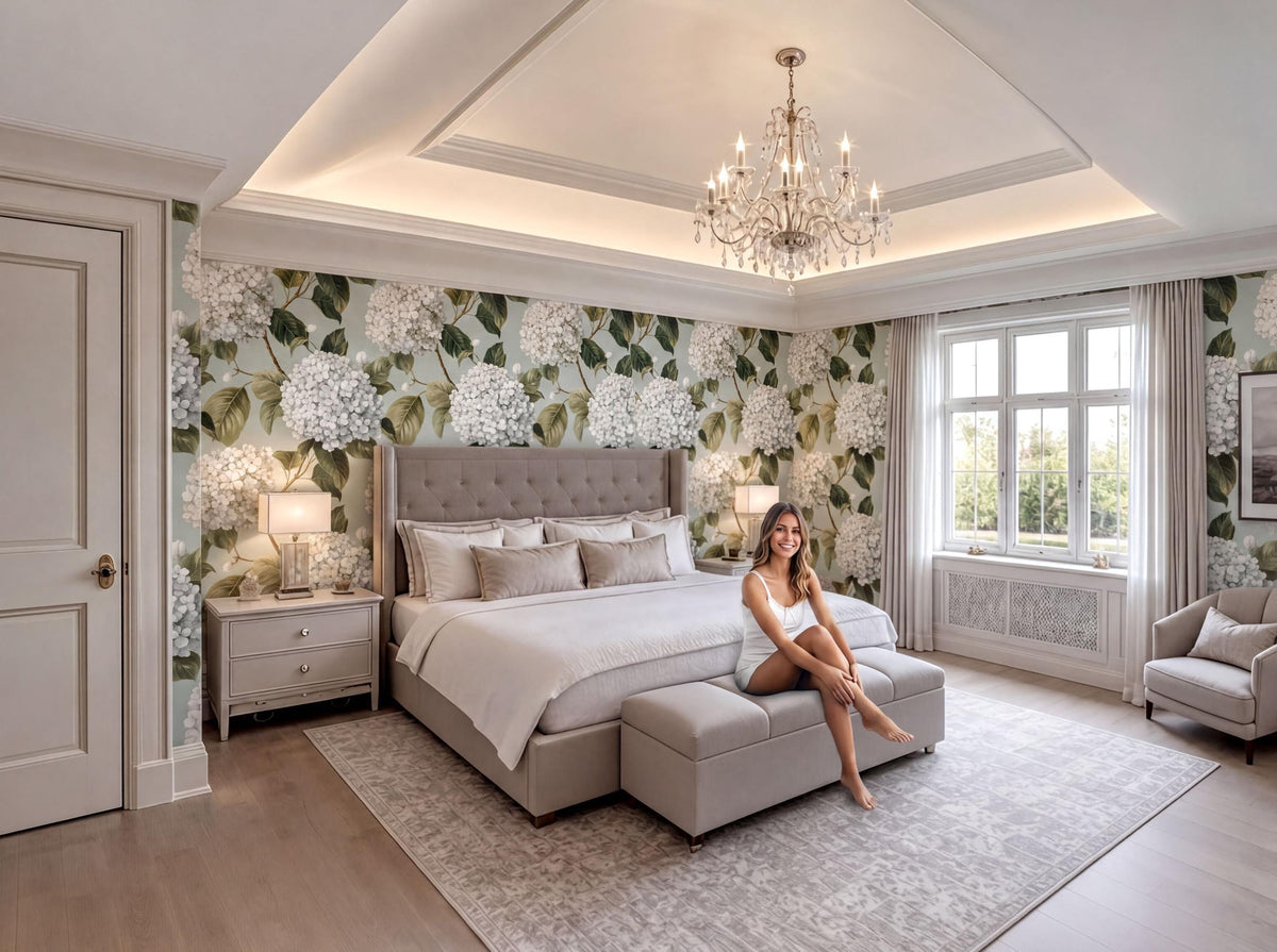 A woman sits on a bench in a luxurious bedroom with a large bed, a chandelier, and a floral wallpaper.
