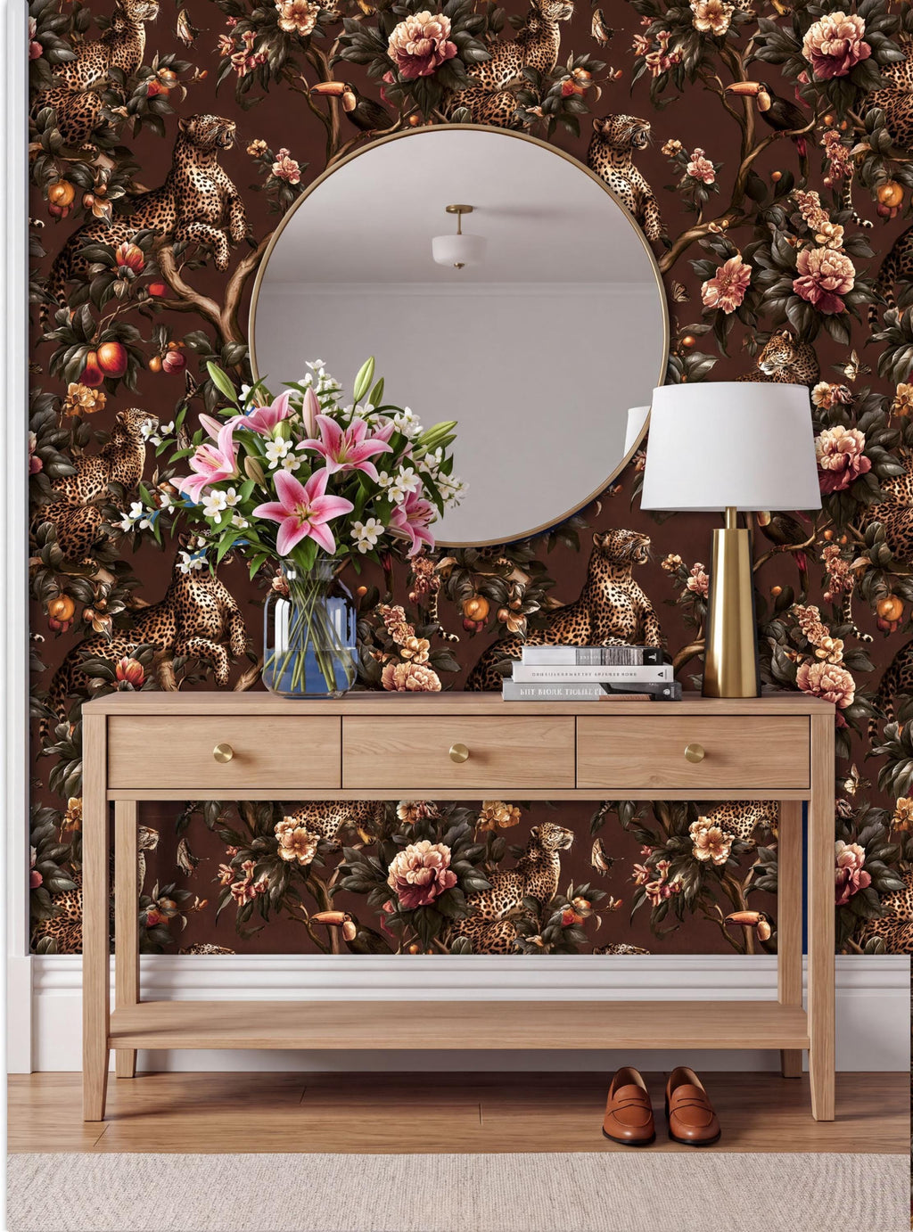 a wooden console table with a round mirror above it, set against a wall with a floral wallpaper. The table has a vase of pink flowers and a lamp on it.