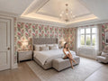 A woman sits on a bench in a large, elegantly decorated bedroom with floral wallpaper, a chandelier, and a large bed.