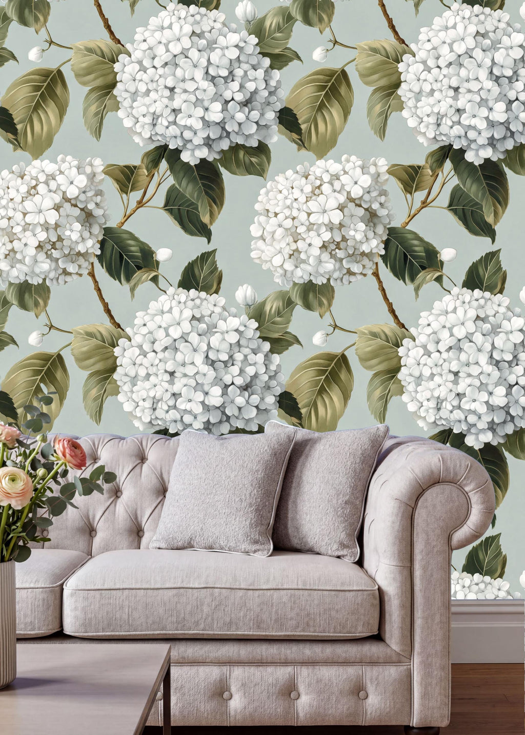 a living room with a white sofa and a vase of flowers on a table. The sofa is positioned against a wall with a floral wallpaper pattern of white hydrangeas and green leaves.