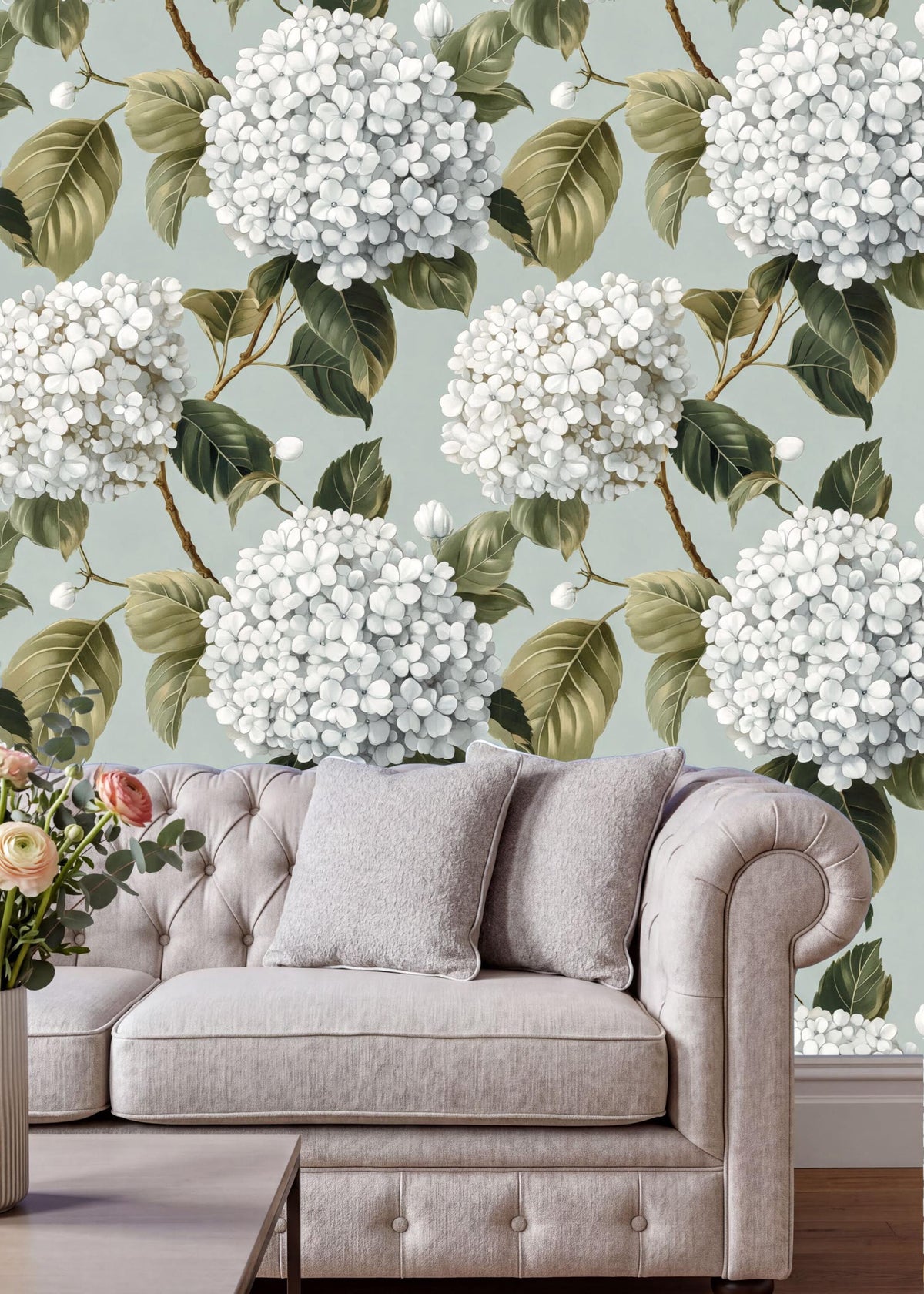 a living room with a white sofa and a vase of flowers on a table. The sofa is positioned against a wall with a floral wallpaper pattern of white hydrangeas and green leaves.