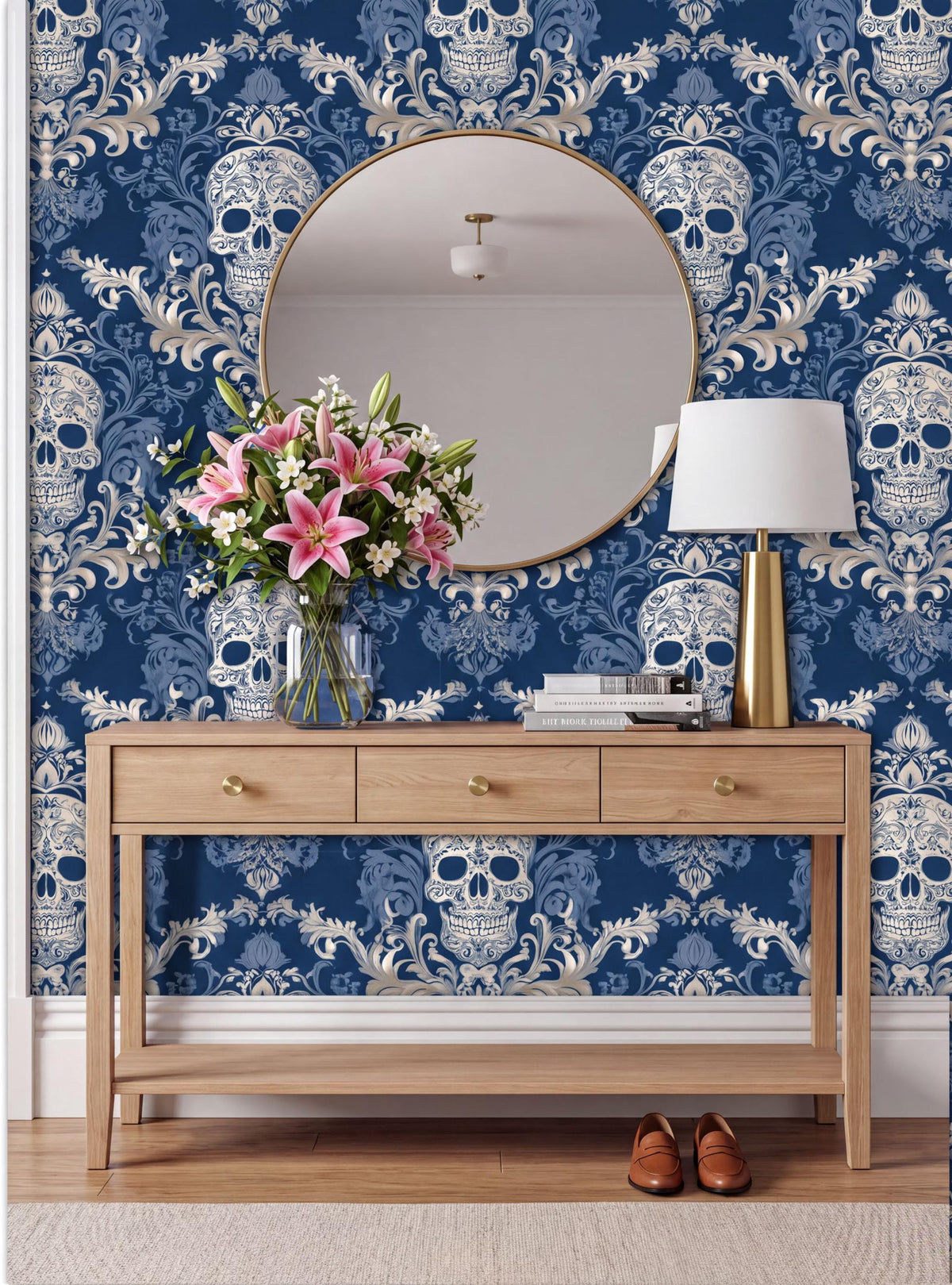 a wooden console table with a round mirror above it, set against a blue and white damask wallpaper with a skull pattern. The table has a vase of pink flowers and a lamp on it.