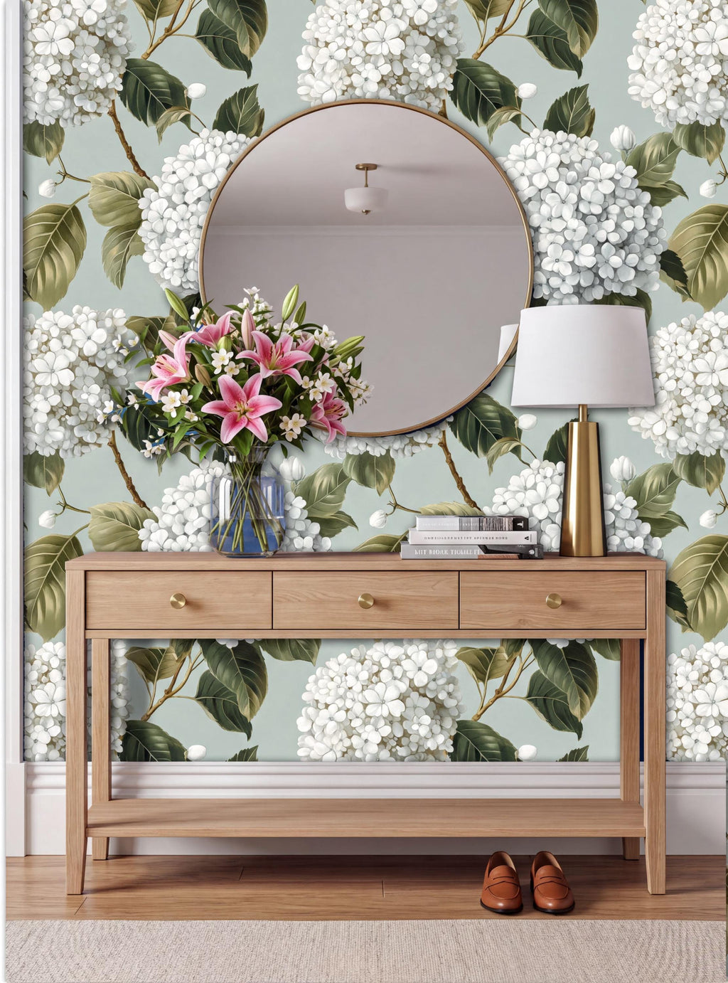 a wooden console table with a round mirror above it, set against a wall with a floral wallpaper. The table has a vase of pink flowers and a lamp on it.