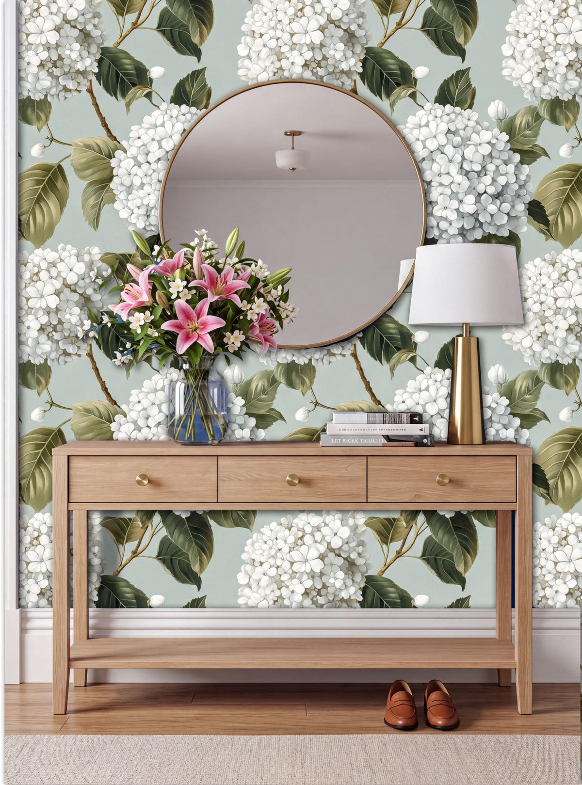 a wooden console table with a round mirror above it, set against a wall with a floral wallpaper. The table has a vase of pink flowers and a lamp on it.