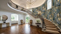 a grand, luxurious entryway with a curved staircase, ornate railings, and a large chandelier overhead. The walls are adorned with a floral wallpaper, and the wooden floor adds to the elegance of the space. The room is filled with natural light from the windows, creating a warm and inviting atmosphere.