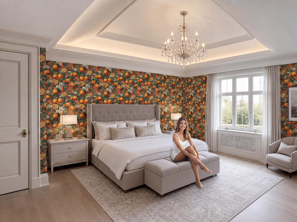 A woman sits on a bench in a large, well-decorated bedroom with a chandelier overhead, orange and green wallpaper, and a large bed with a tufted headboard.
