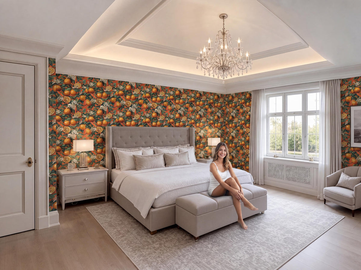 A woman sits on a bench in a large, well-decorated bedroom with a chandelier overhead, orange and green wallpaper, and a large bed with a tufted headboard.