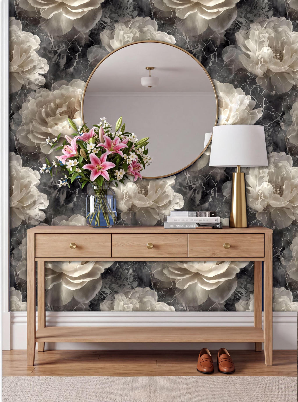 a wooden console table with a round mirror above it, a vase of pink flowers, and a lamp on the table. The wall behind the table has a floral wallpaper design.