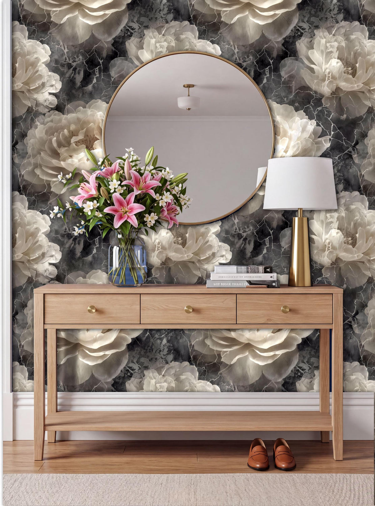 a wooden console table with a round mirror above it, a vase of pink flowers, and a lamp on the table. The wall behind the table has a floral wallpaper design.