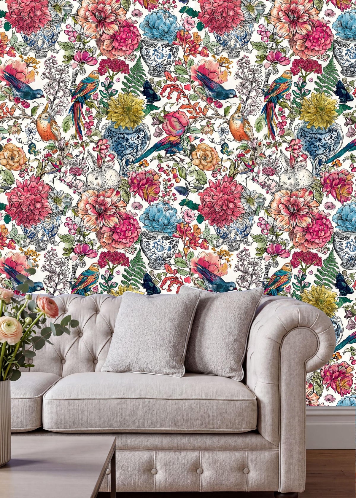 a vibrant floral wallpaper with various colorful flowers and birds, creating a lively and cheerful atmosphere. In the foreground, there is a white tufted sofa with a gray throw pillow, and a vase of flowers on the floor.