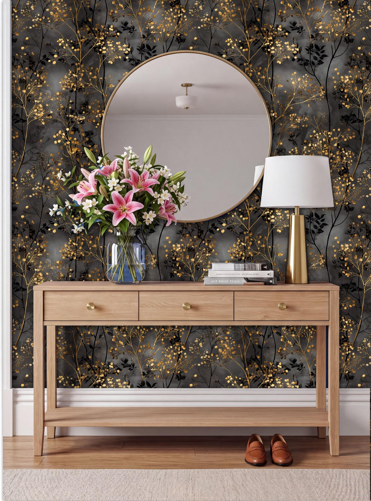 a wooden console table with a round mirror above it, a vase of pink lilies, and a lamp on the table. The wall behind the table has a black and gold floral wallpaper.