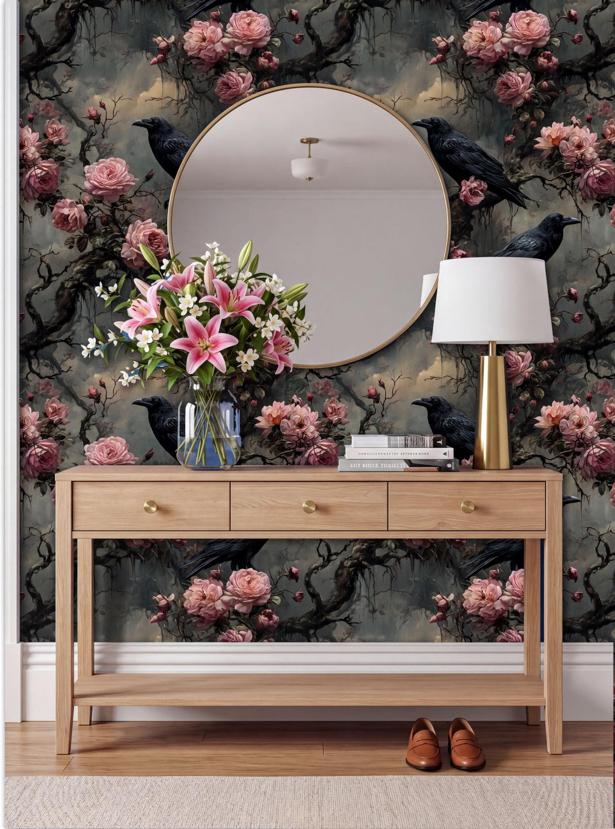 a wooden console table with a round mirror above it, set against a wall with a floral wallpaper. The table has a vase of pink flowers, a lamp, and a pair of shoes placed on it.