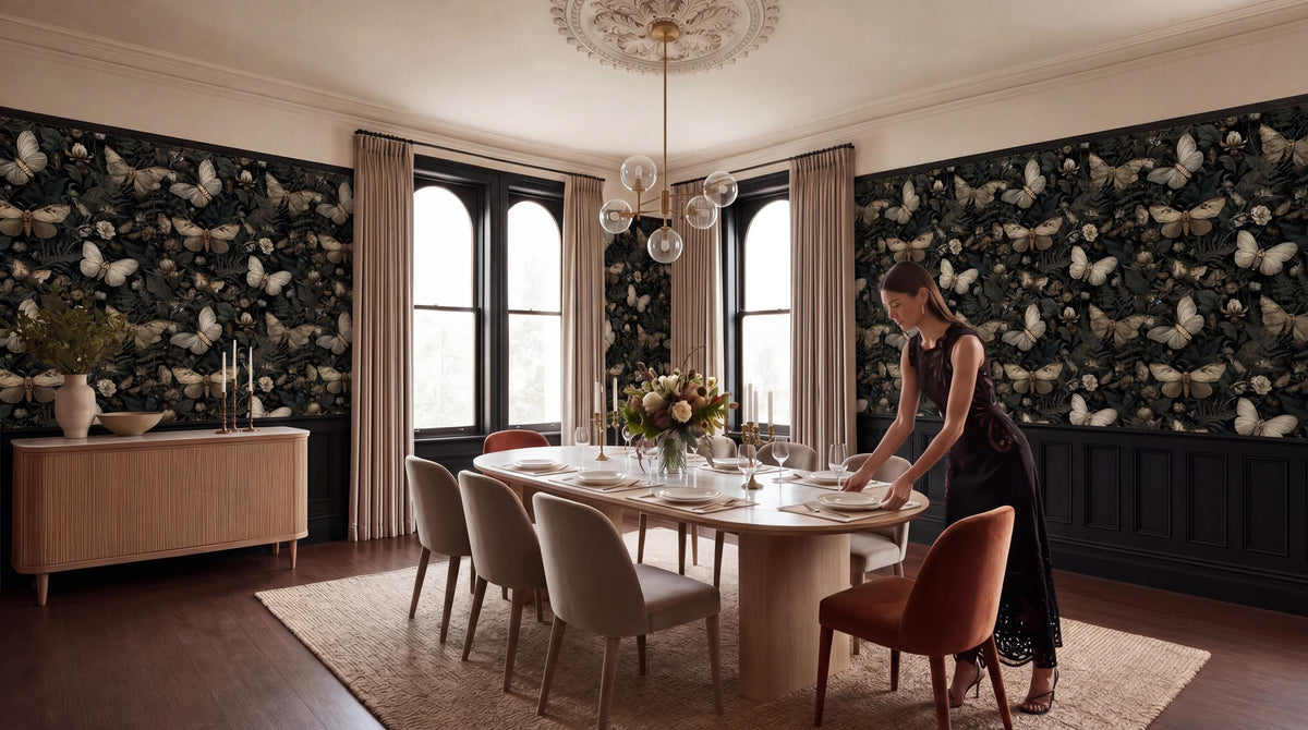 A woman in a black dress is setting the table in a dining room with a large, ornate chandelier overhead. The room features a black and white butterfly wallpaper, a wooden dining table with white plates and silverware, and a large window with curtains.
